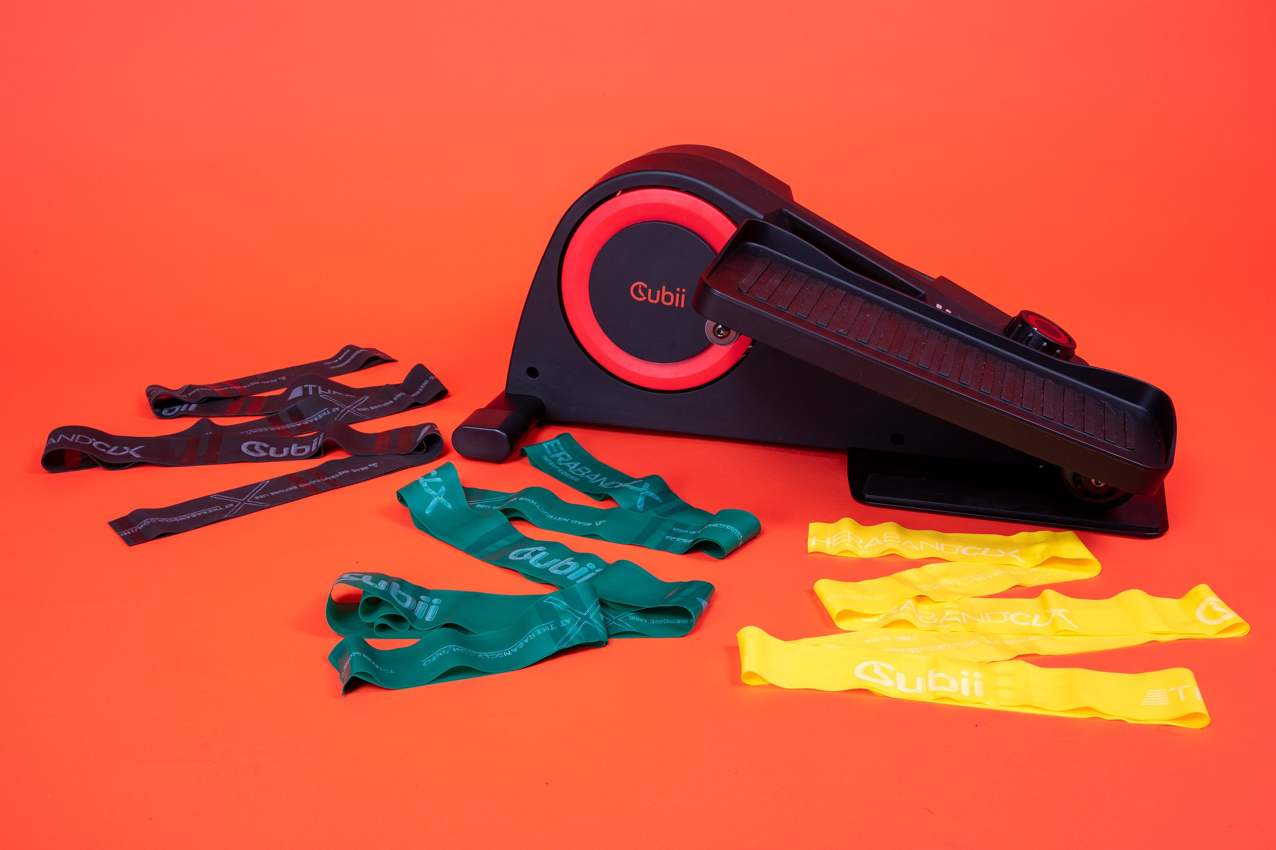 A black and red exercise treadmill with colorful resistance bands in black, green, and yellow laid out on an orange background.