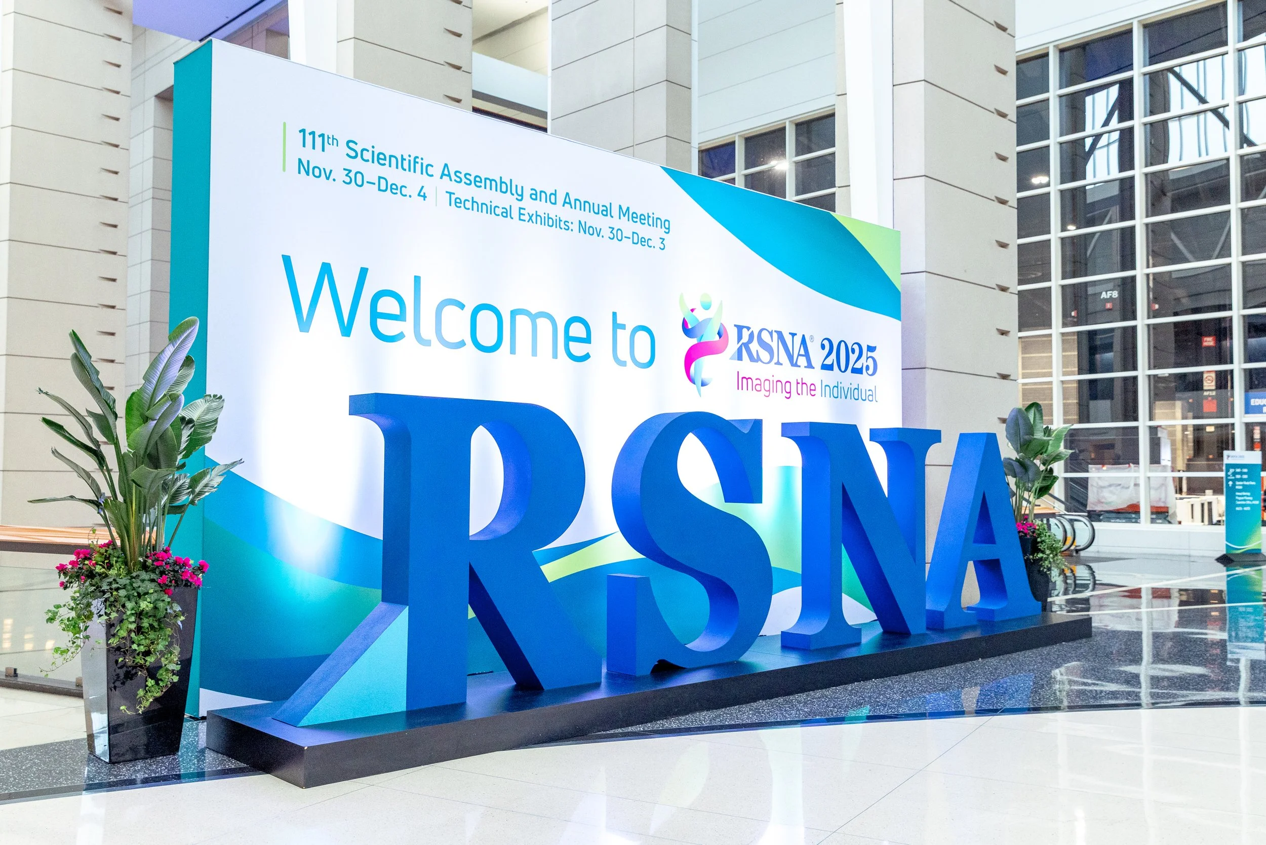 Large welcome sign for the 111th Scientific Assembly and Annual Meeting of RSNA, taking place from November 30 to December 4, with technical exhibits from November 30 to December 3. The sign features colorful logos and text, with decorative plants at