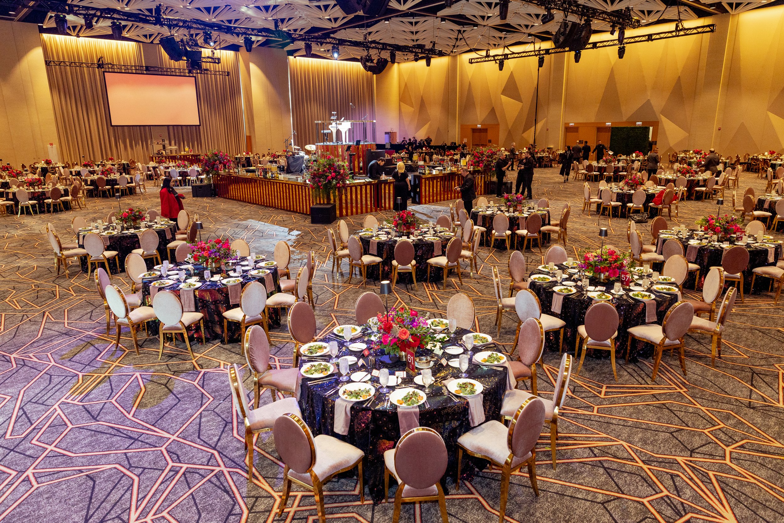 A large banquet hall set up for an event with numerous round tables covered with black tablecloths, each adorned with a centerpiece of pink and red flowers. The tables are set with plates, utensils, and salad bowls. There is a stage in the background