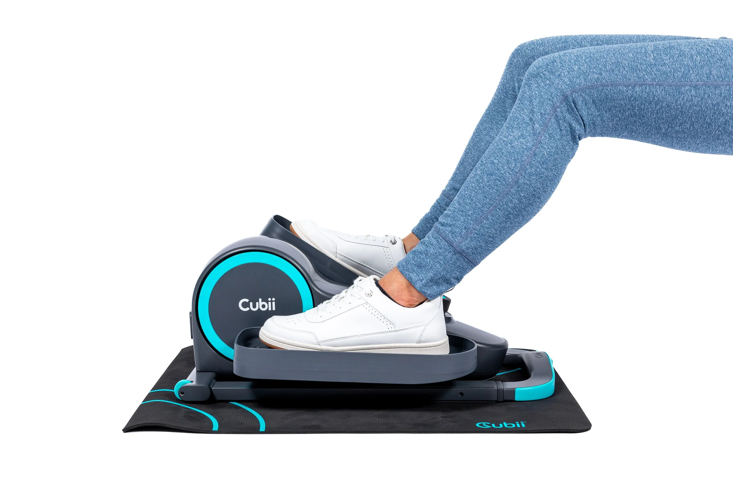 A person wearing blue jeans and white sneakers is using a Cubii under-desk elliptical machine placed on a black workout mat.