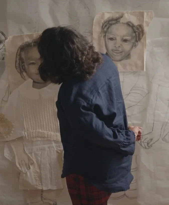 A woman with dark curly hair and a dark jacket working on two detailed pencil sketches of young girls on large piece of fabric on a wall.