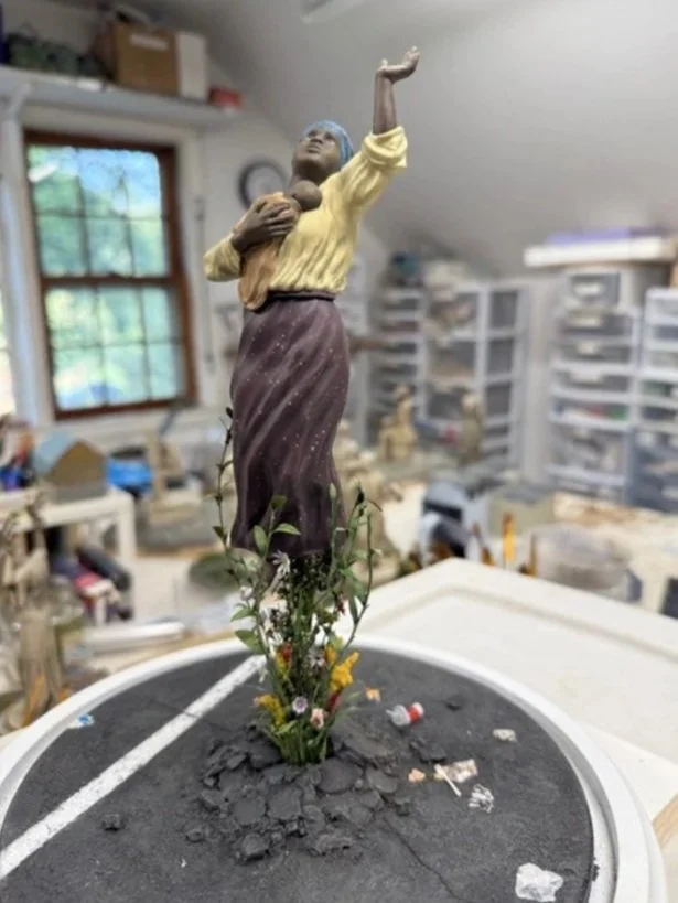 A religious statue of a woman with her arm raised and holding a child, set on a circular base with earth and small plants, in a studio with storage bins and a window.