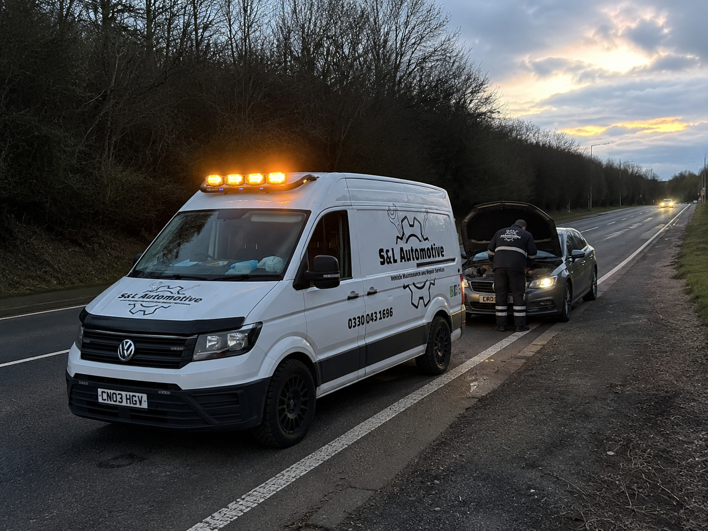 A vehicle broken down on the side of a highway at dusk with a mechanic inspecting the engine, and a S&L Automotive service van parked nearby with its lights on.