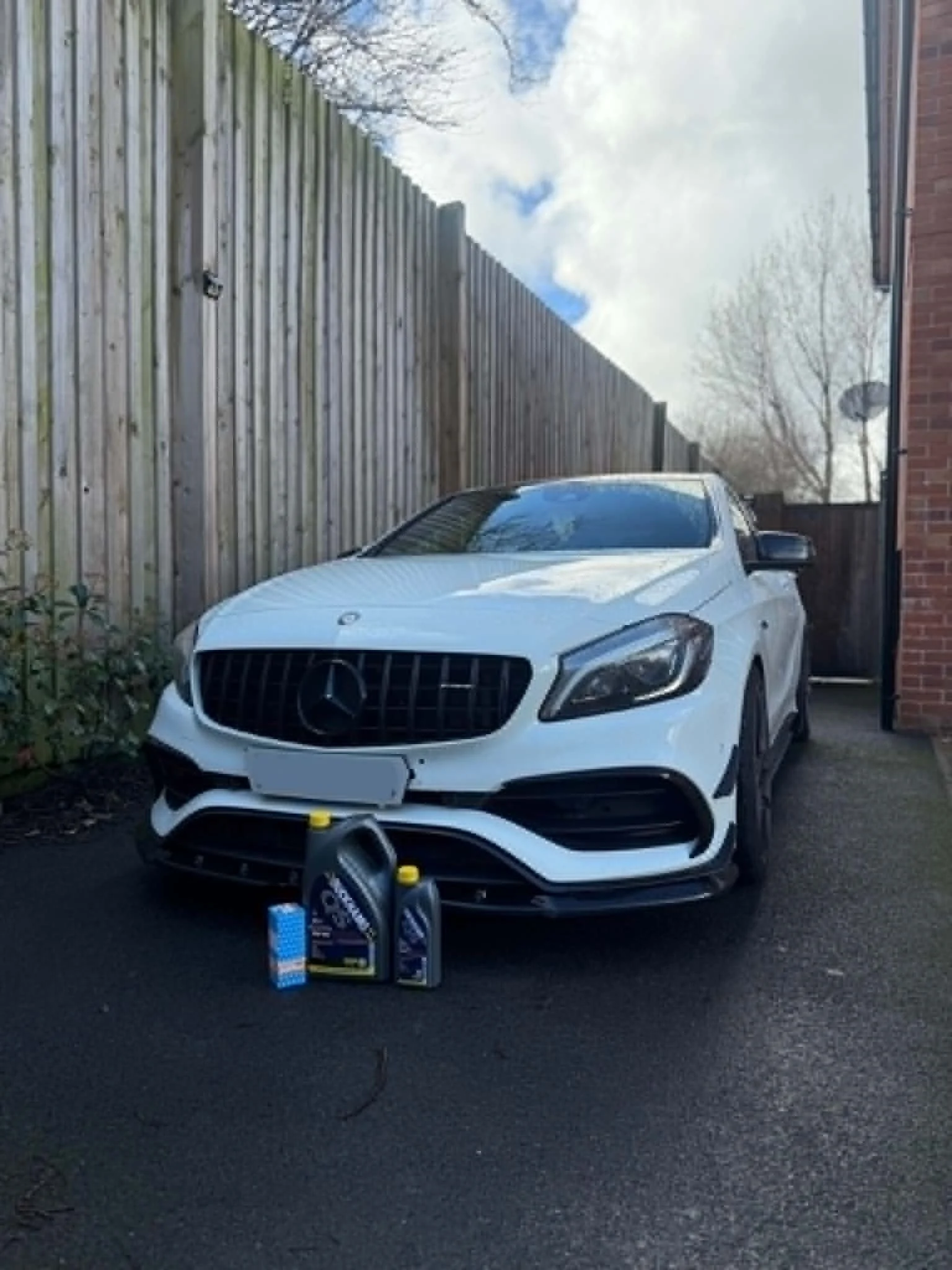 White Mercedes-Benz car parked in a driveway in front of a wooden fence and brick house wall, with some open car parts and tools on the ground nearby.