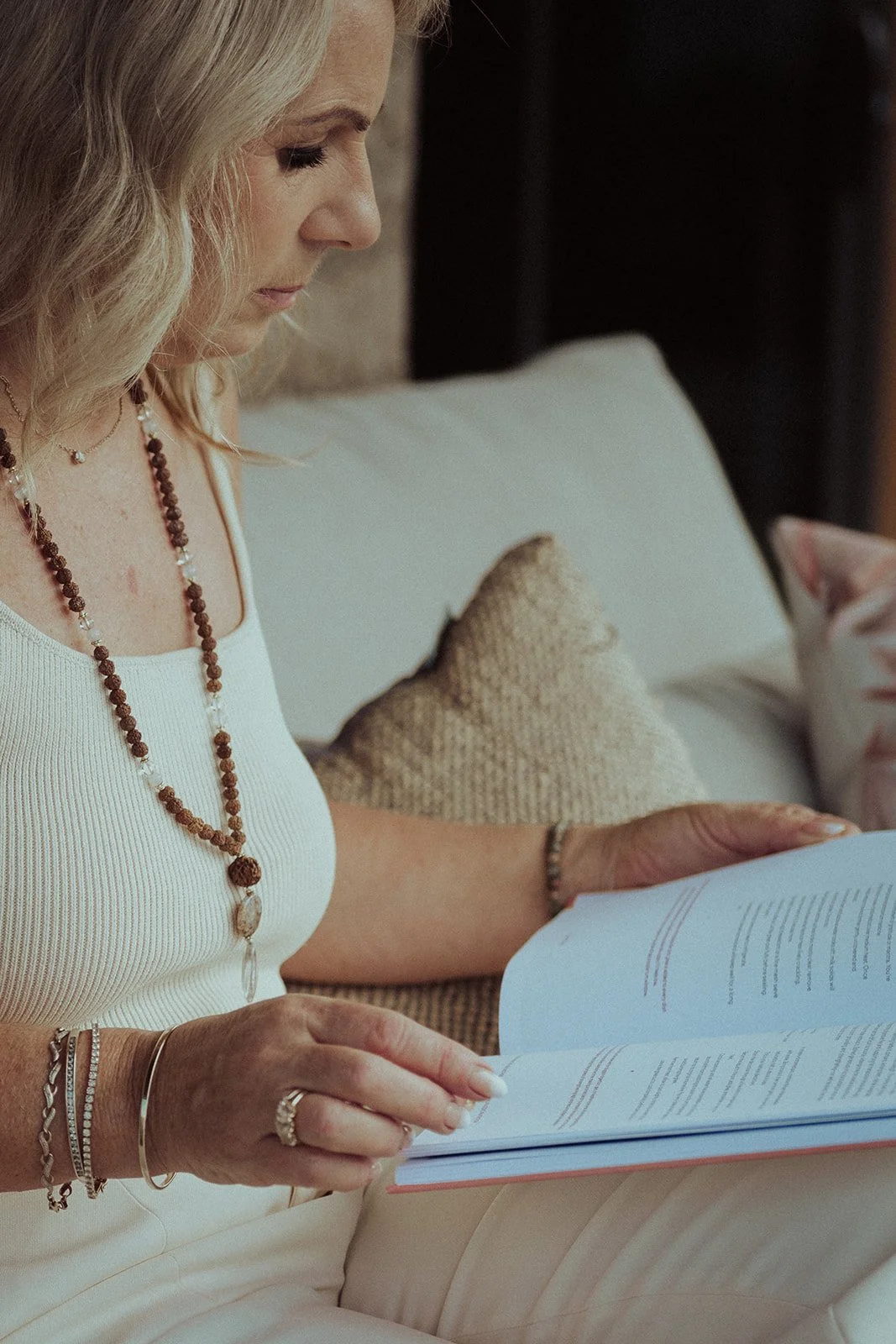 Woman with blonde hair wearing necklaces and bracelets, reading a book or magazine on a beige couch.