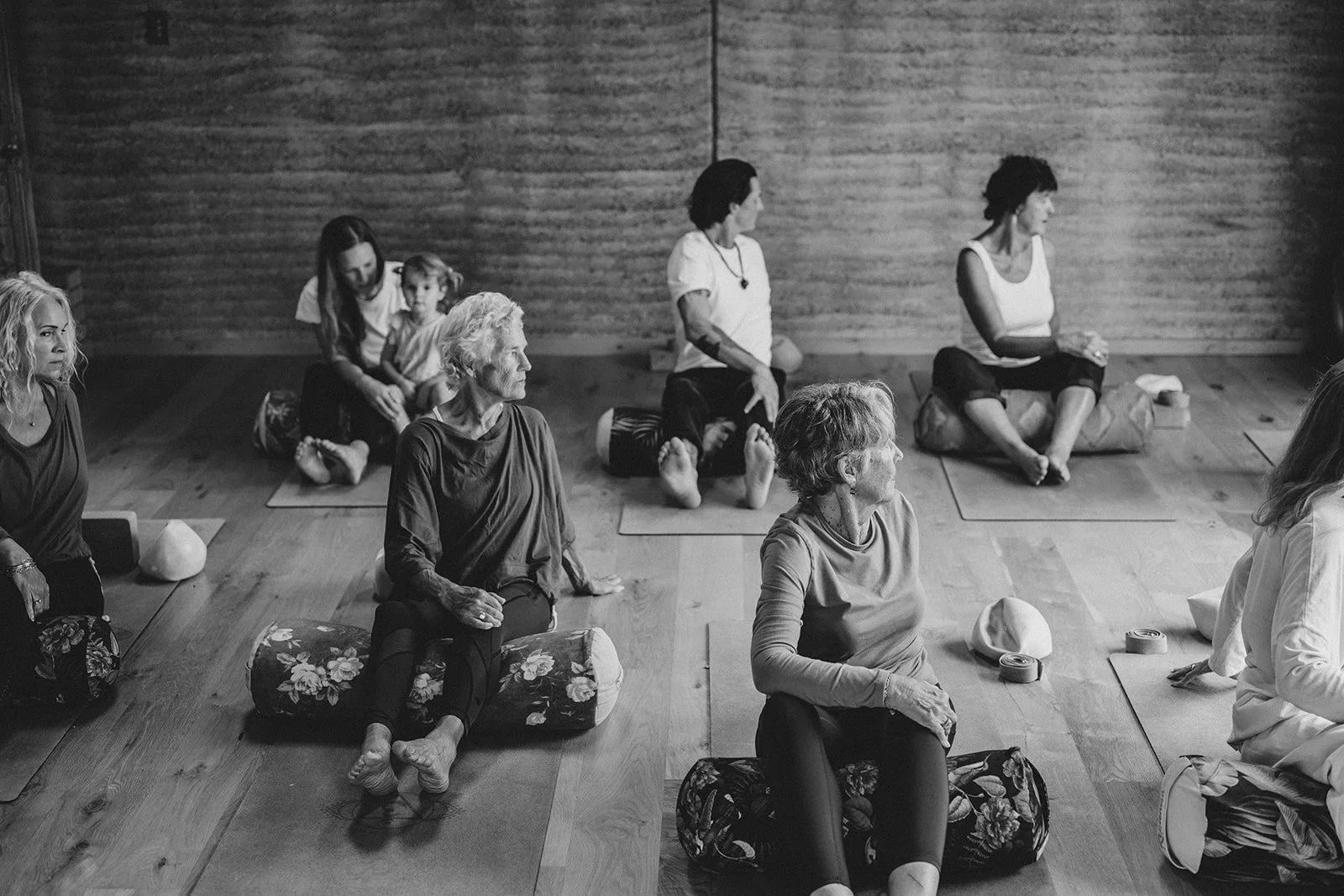 Group of elderly and middle-aged women attending a yoga or meditation class in a room with wooden walls, sitting on mats with folded arms, looking attentive, with some children present in the background.