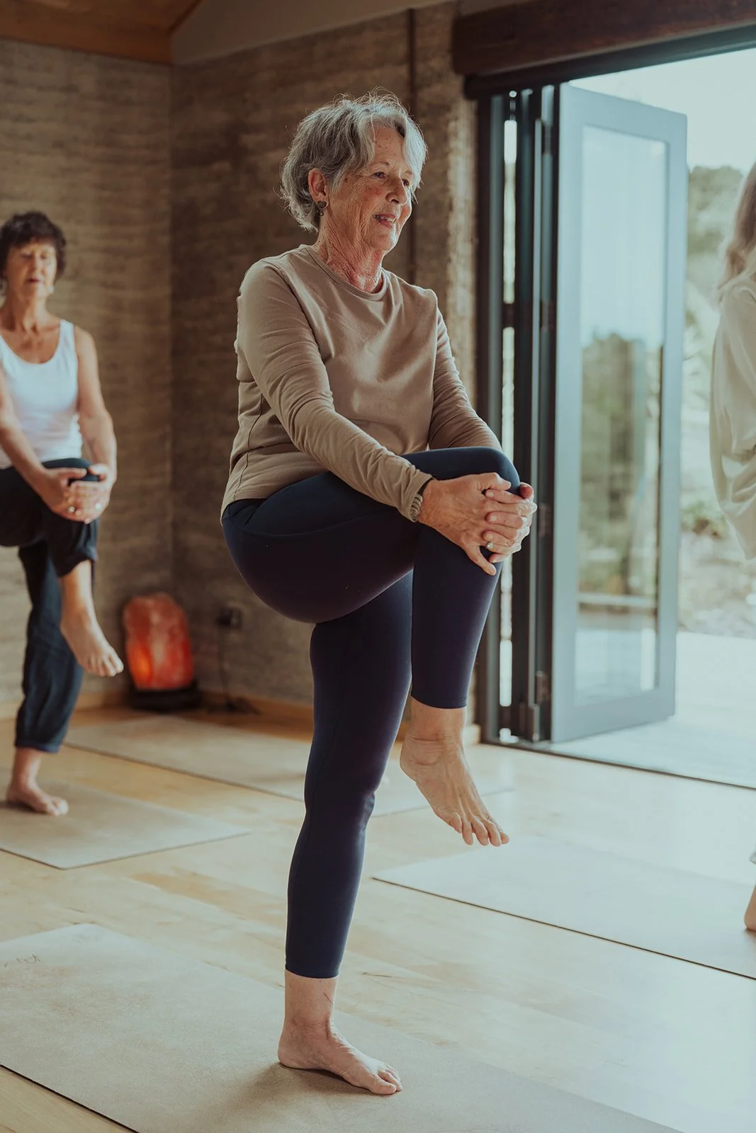 Older woman practicing yoga in a studio, balancing on one leg with a knee held to her chest. Others are in the background, also participating in a yoga class.