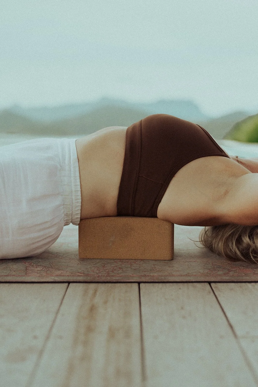 Person lying on their back with a yoga block under their neck during yoga practice on a wooden floor with a mountain view in the background.
