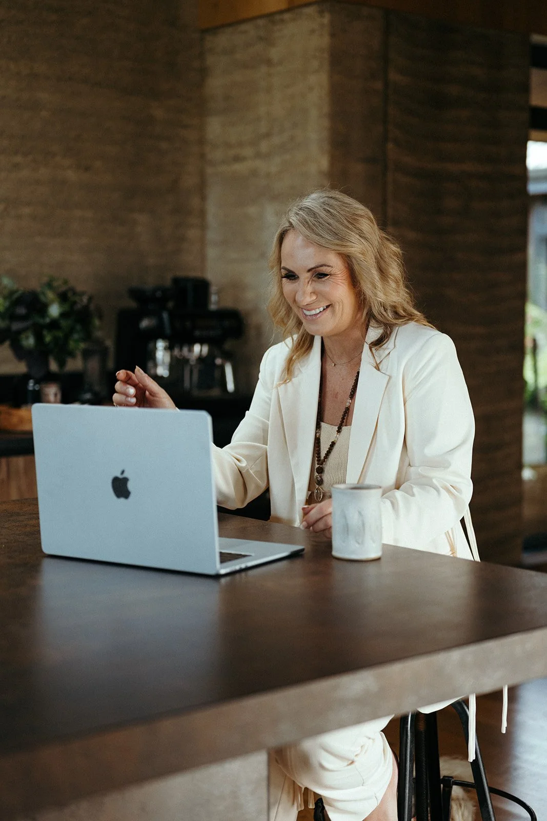 Smiling woman in white blazer sitting at a wooden table with a MacBook and coffee mug, working in a cozy cafe.