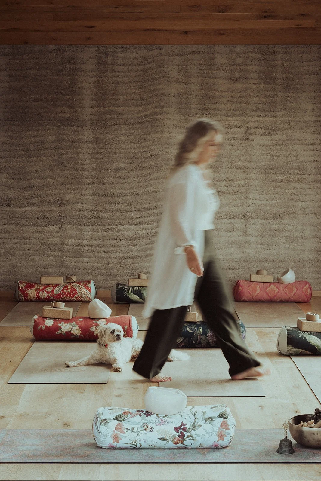 A person walking barefoot on a yoga mat with a dog lying next to them, in a room with a wooden floor and a textured wall, with yoga bolsters and props in the background.