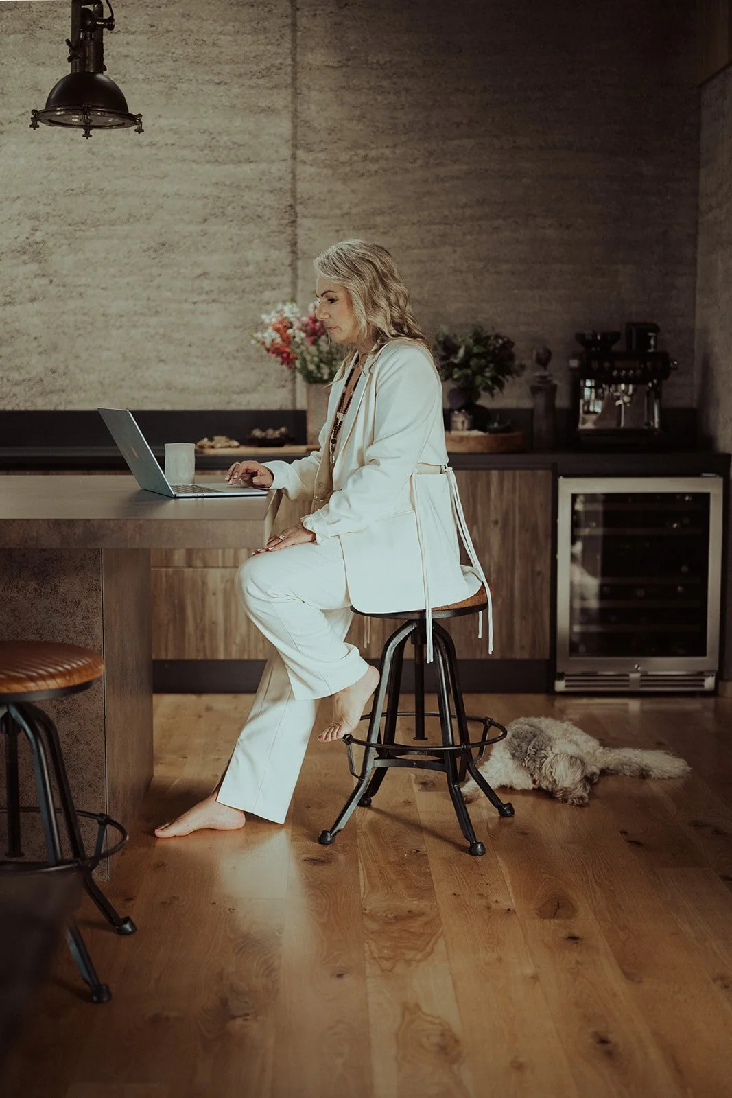 A woman with blonde hair in a white suit sits at a kitchen counter using a laptop, with a dog lying on the floor nearby and flowers in the background.