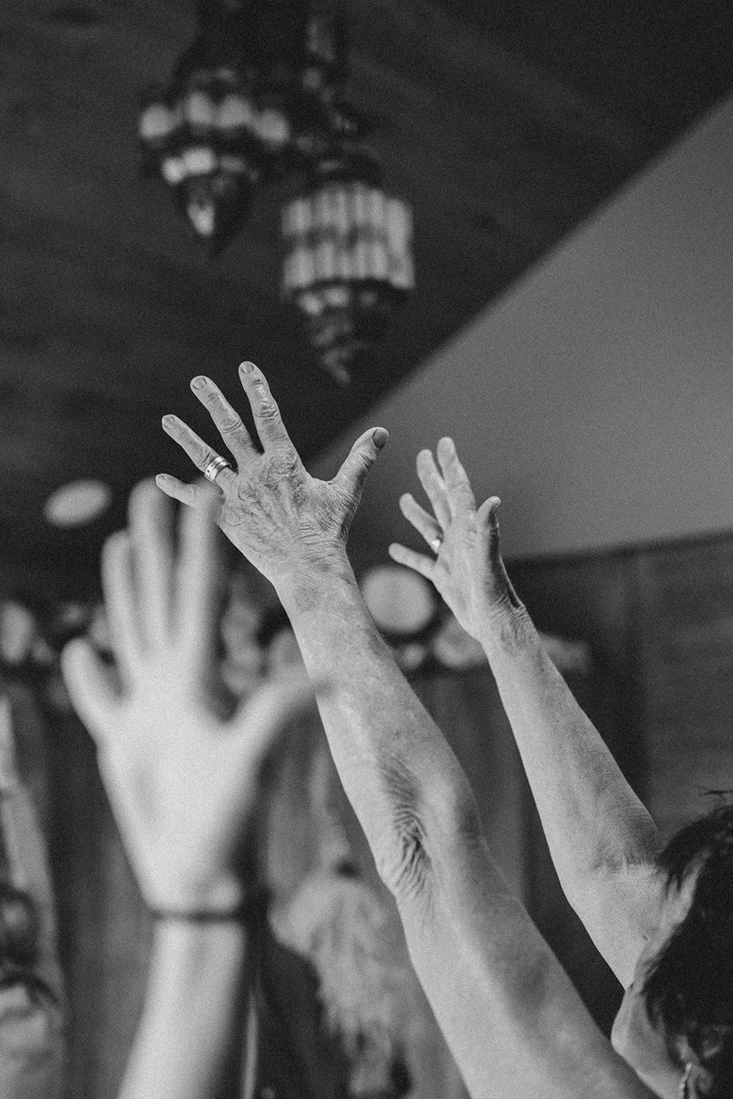 Black and white photo of multiple hands reaching upwards, some with rings, in front of a dark ceiling with chandeliers.