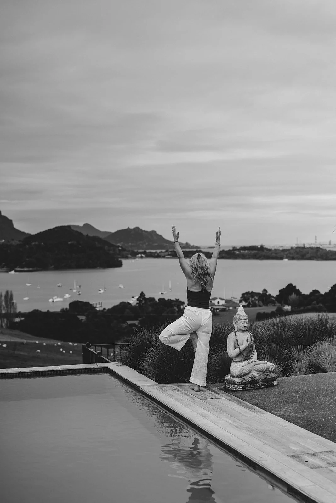 A woman practicing yoga outdoors on a deck near water, with a scenic lake, boats, mountains, and a cloudy sky in the background, and a Buddha statue nearby, in black and white.