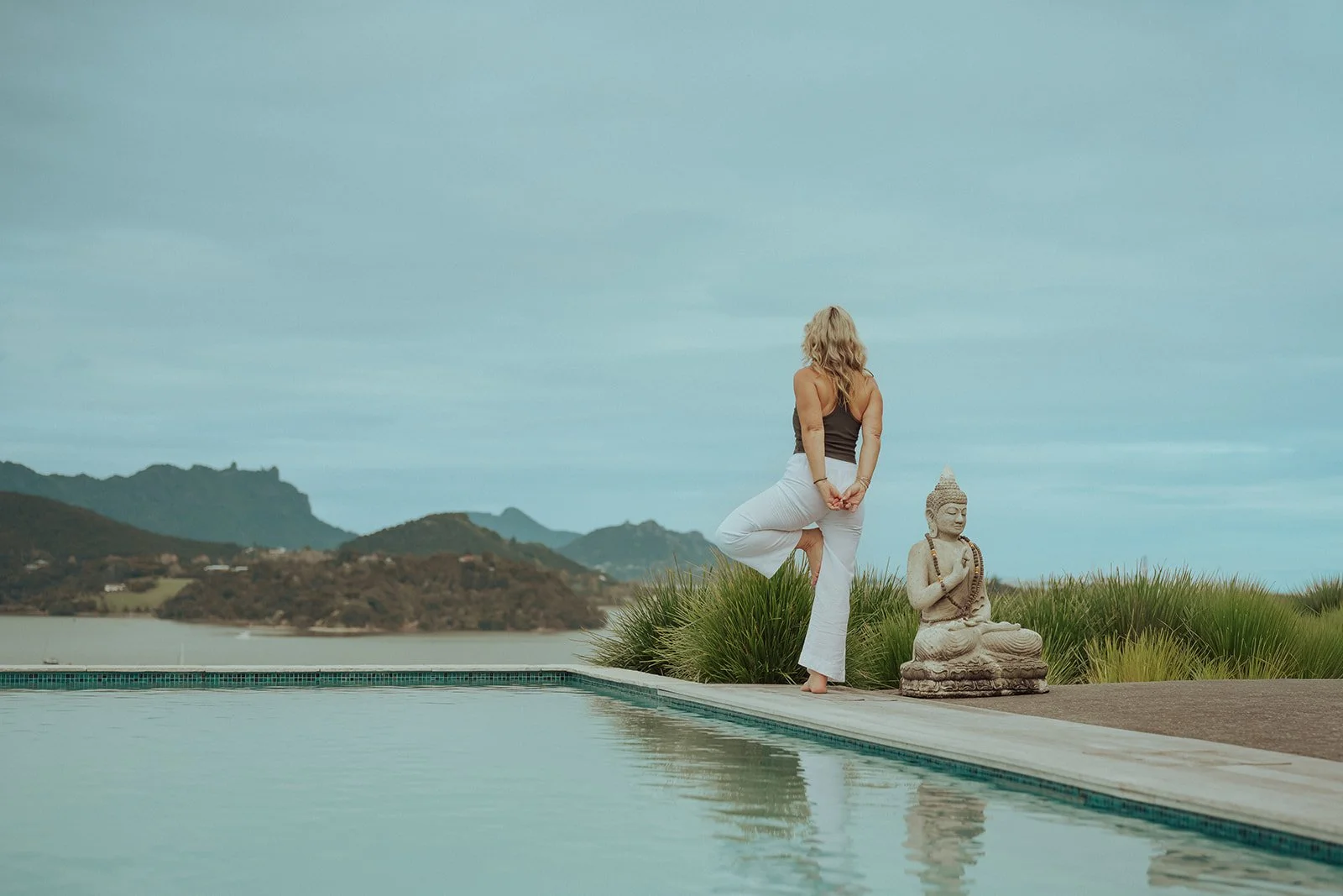 Woman practicing yoga outdoors near a pool with a statue of Buddha and a scenic hillside in the background