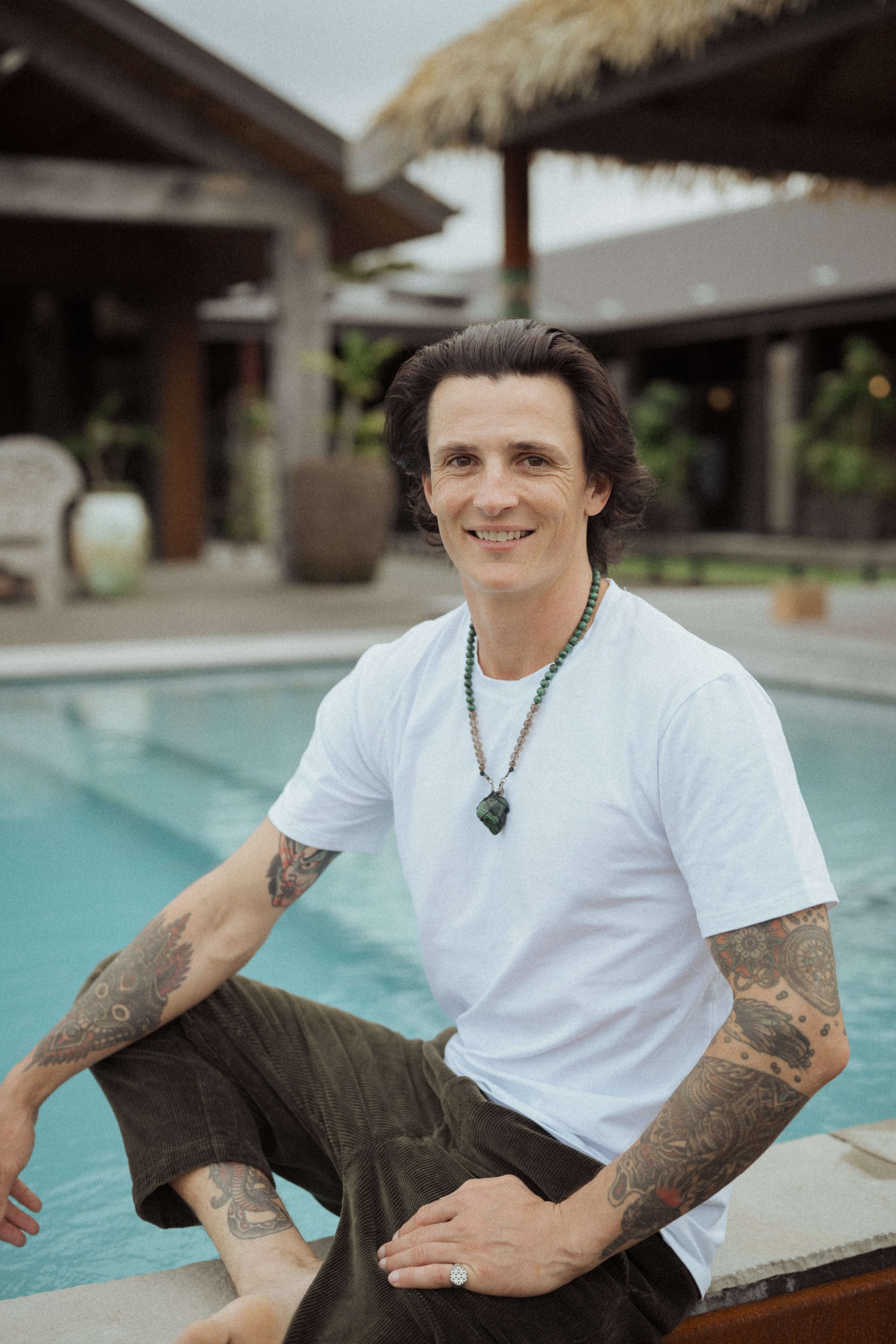 A man with dark hair and tattoos on his arms wearing a white t-shirt and beaded necklaces sitting by a pool.