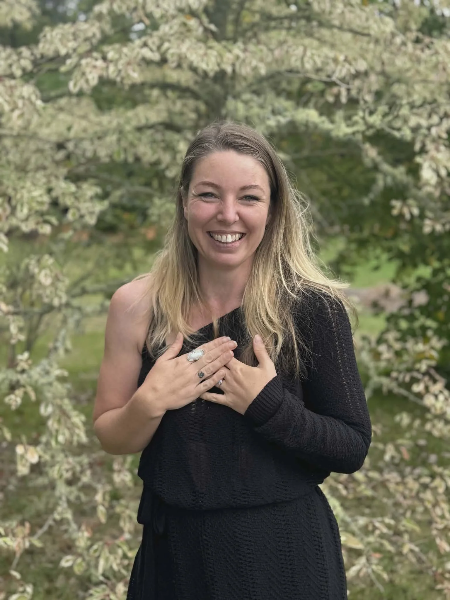 A woman with blonde hair smiling outdoors in front of a flowering tree, wearing a black sweater and ring.