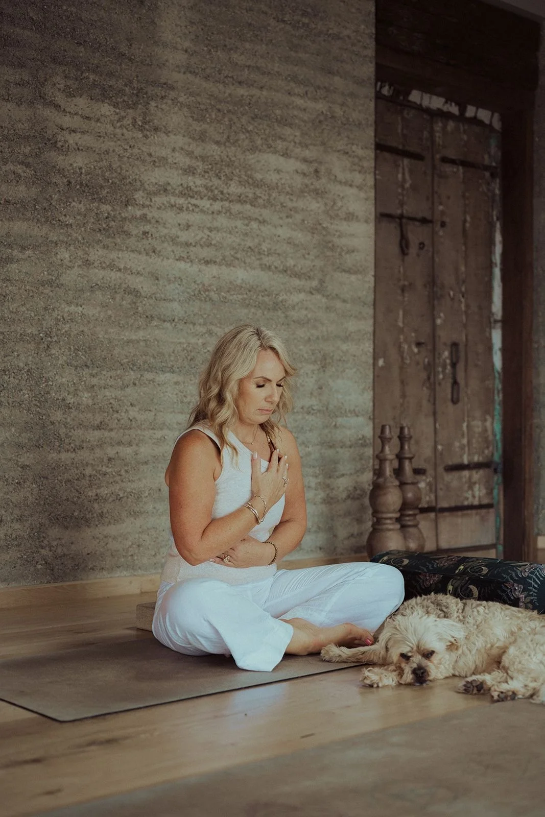 A woman with blonde wavy hair in white clothing sitting on a yoga mat with her eyes closed, one hand on her chest and the other on her stomach, next to a sleeping light-colored dog on the floor, in a rustic room with a textured wall and wooden door.