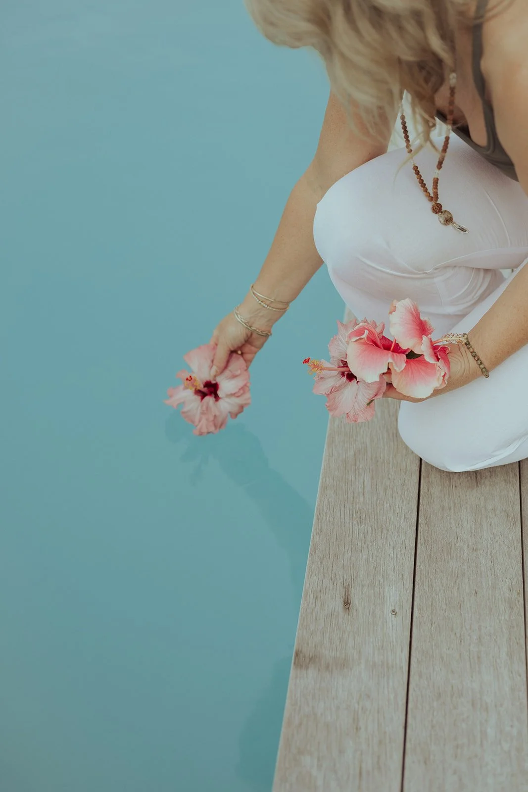 A woman in white pants holding pink hibiscus flowers while sitting on a wooden dock beside a blue body of water.