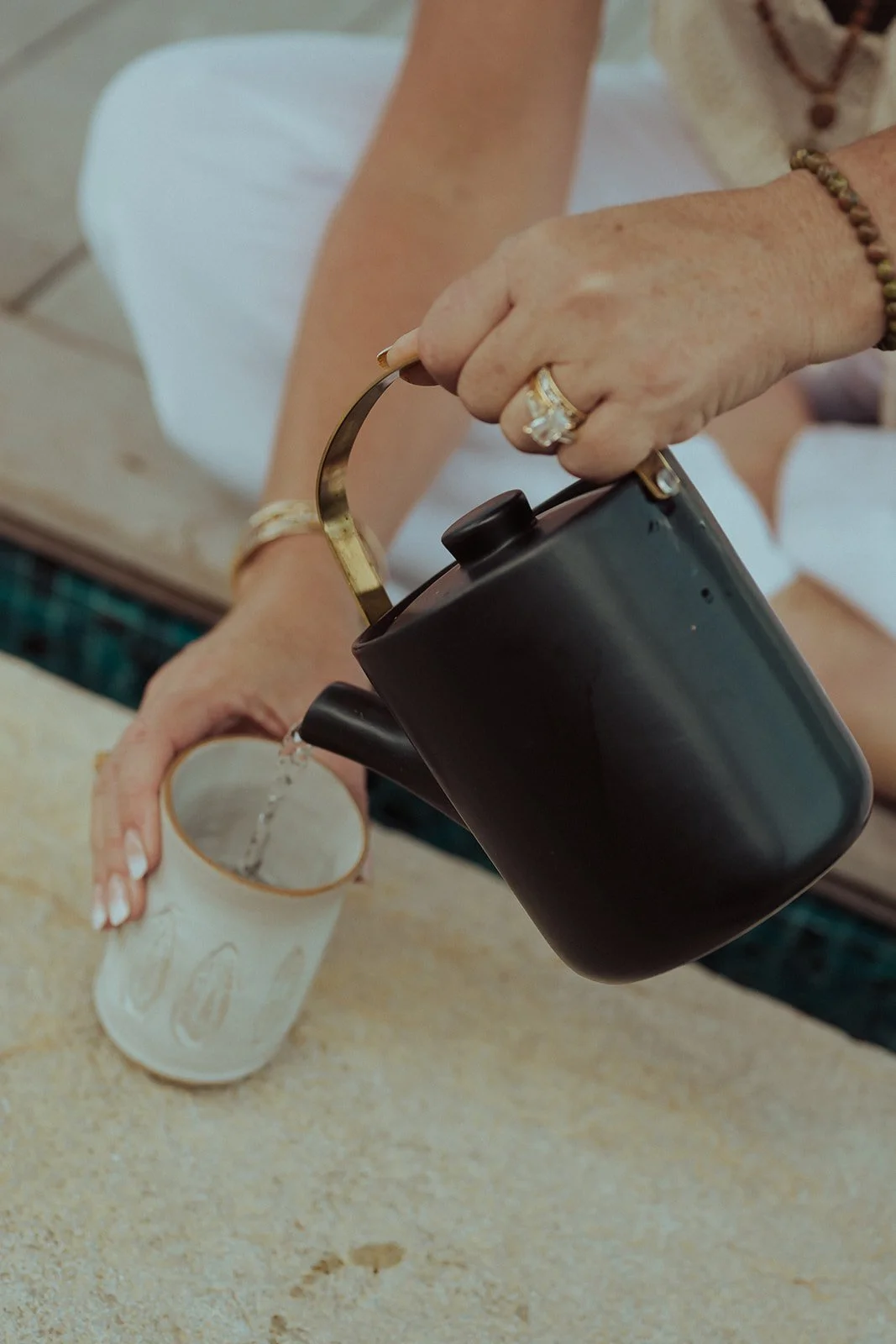 A person is pouring hot water from a black kettle into a ceramic mug on a beige countertop.