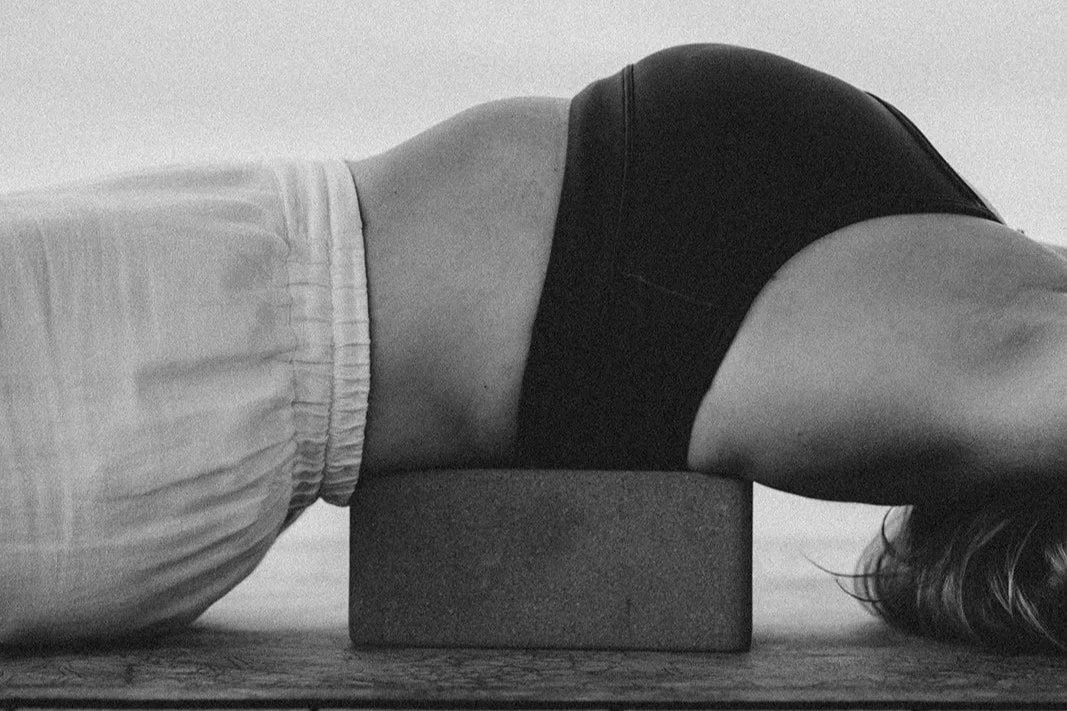 Person demonstrates a yoga pose with their upper back on a yoga block, lying face down on the floor.