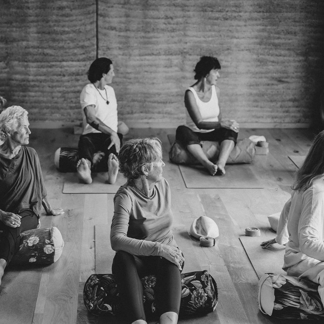 A group of people participating in a yoga or meditation class in a studio with wooden walls. Some are sitting or kneeling on mats, and others are seated on cushions, engaging in a session led by an instructor.