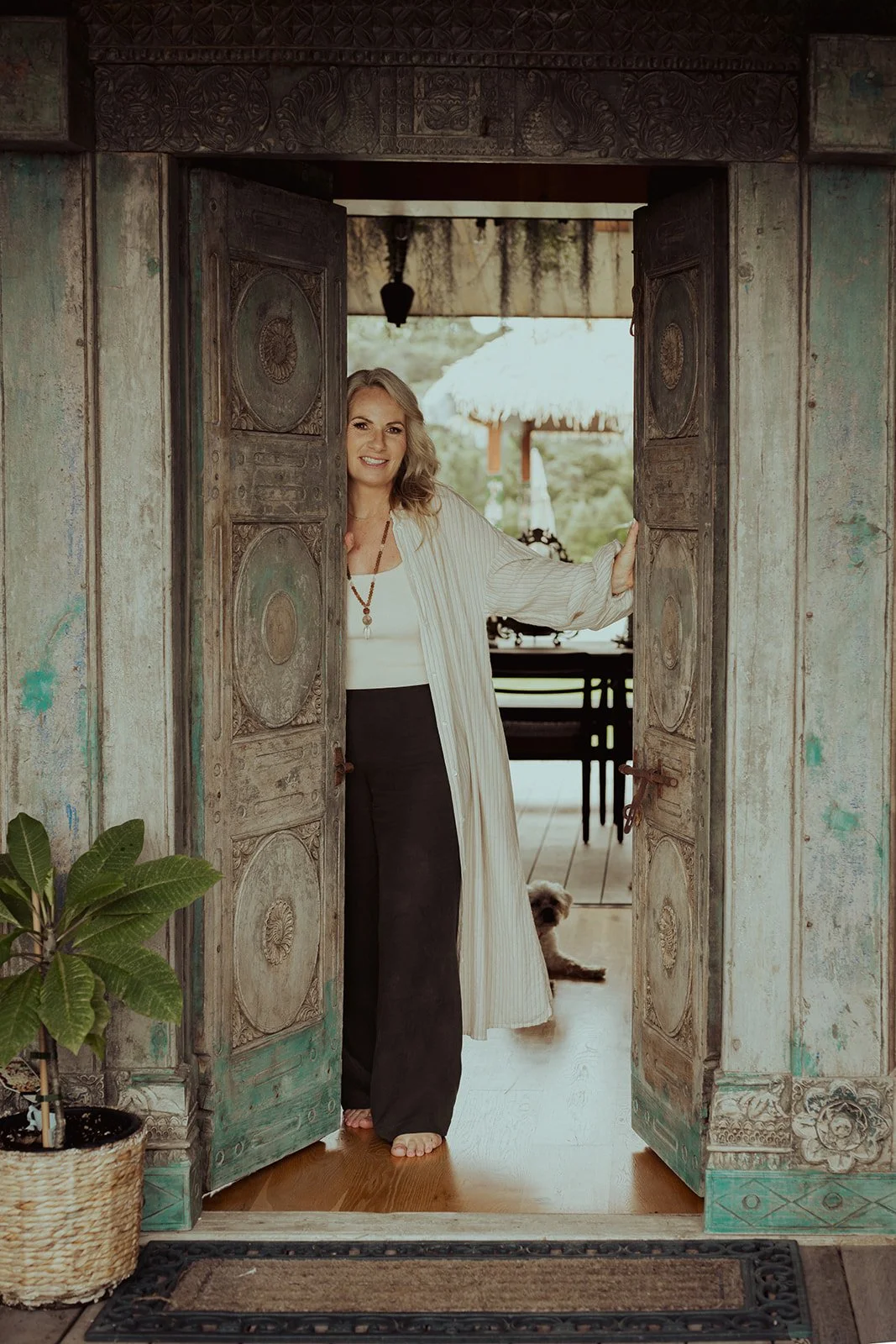 A woman standing in an open, carved wooden doorway with a small dog sitting in the background, on a porch with a thatched roof, greenery, and outdoor furniture.