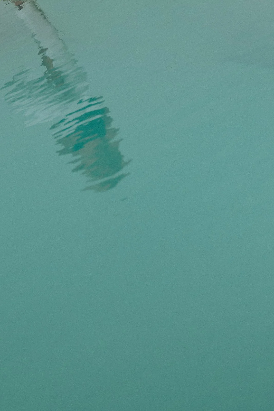 Reflection of a person and possibly a part of a building on the surface of a swimming pool with clear turquoise water.