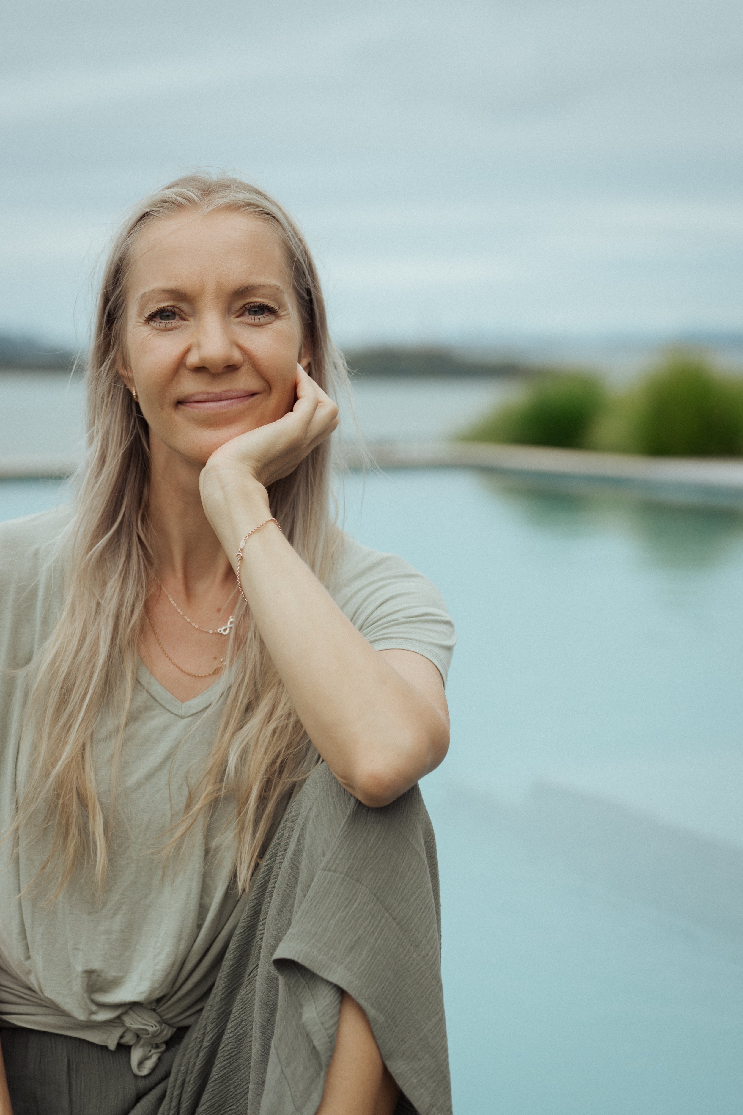 A woman with long blond hair sitting outdoors near a body of water, smiling gently with her hand resting on her cheek, wearing a light-colored top and earrings, with a blurred background of sky and greenery.