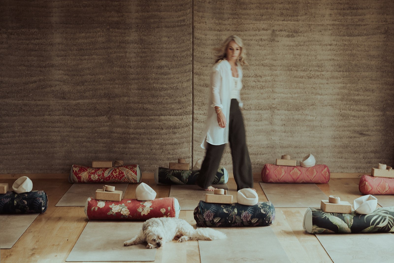 A woman practicing yoga on a mat in a room with meditation cushions and a dog resting on the floor.