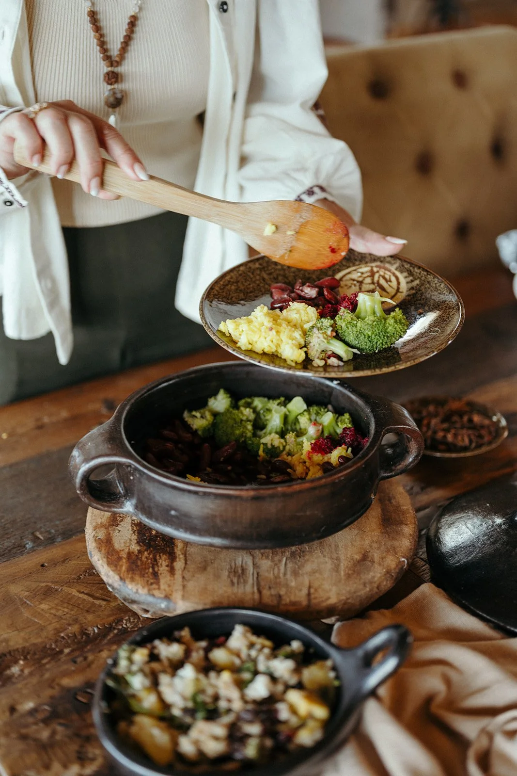 Person serving salad from black pot onto a plate, with broccoli, scrambled eggs, beans, and herbs on a wooden table.
