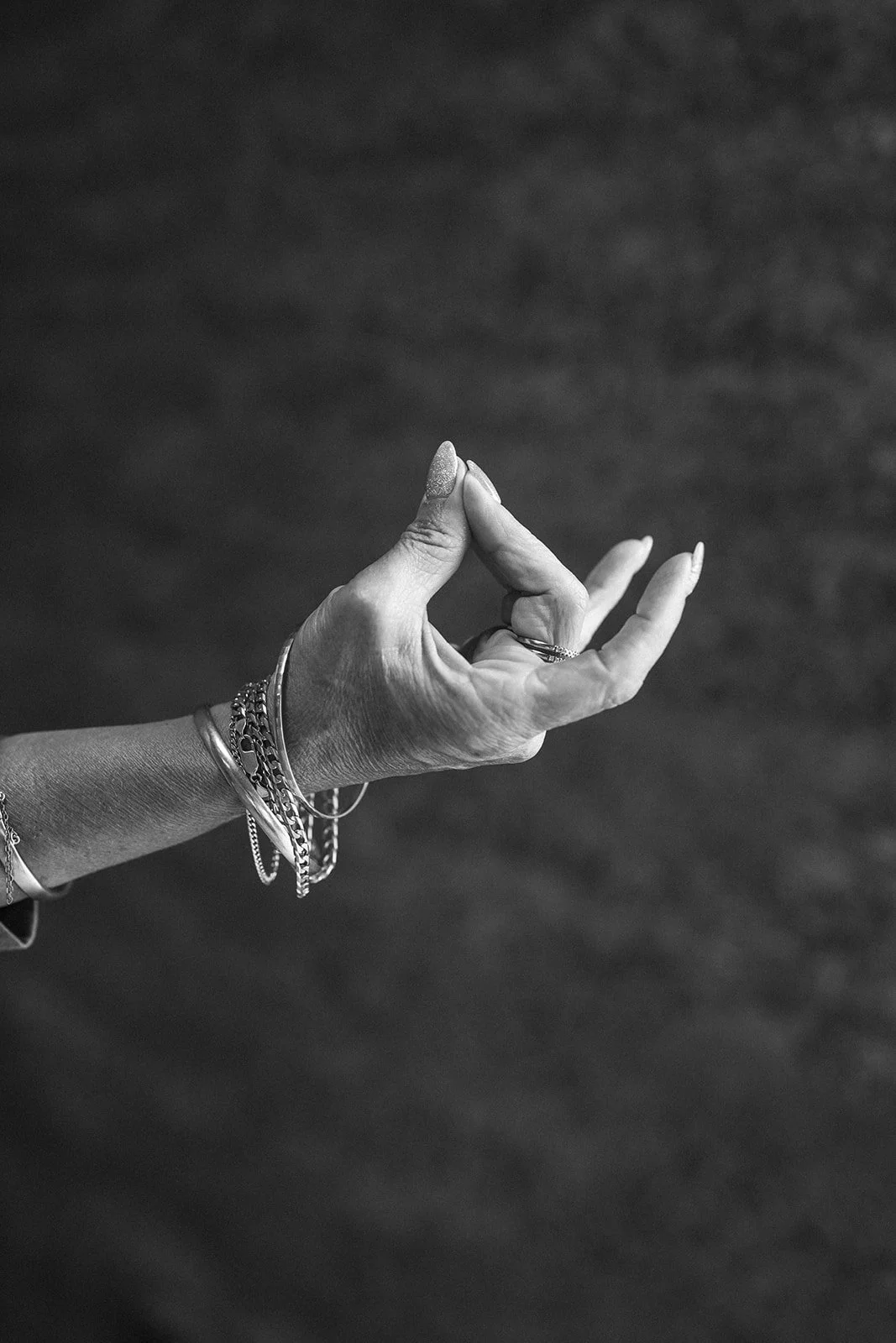 Black and white photo of a woman's hand making a mudra gesture with her thumb and index finger touching, adorned with bracelets and rings.