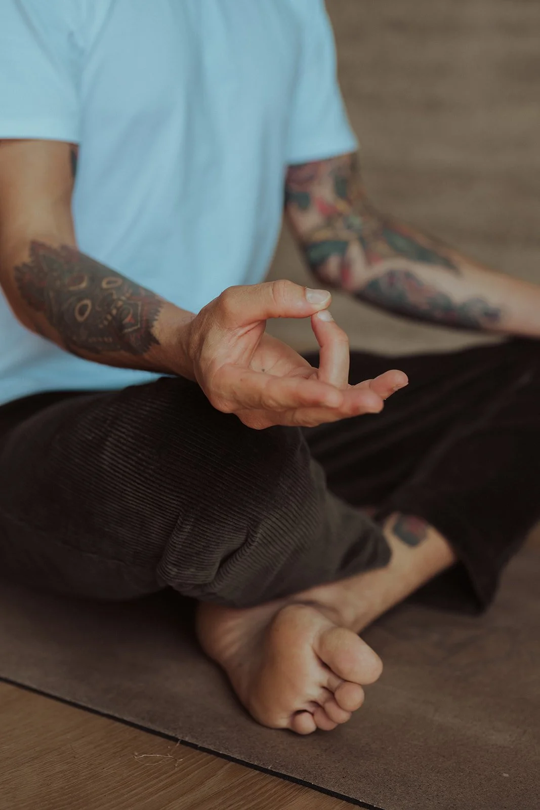 Person sitting on the floor in a cross-legged meditation pose, wearing a light blue shirt and dark pants, with colorful tattoos on their arms.