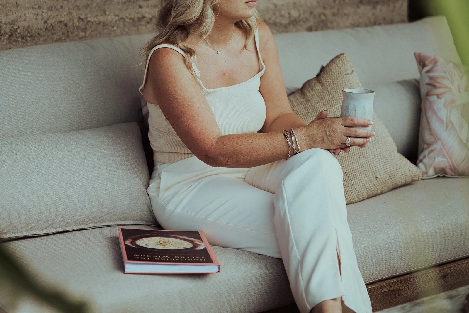 A woman with blonde hair sitting on a beige outdoor sofa, holding a white ceramic mug, with a book on the sofa near her. She is wearing a white sleeveless top and white pants, with jewelry on her wrist and fingers. There are decorative pillows on the sofa.