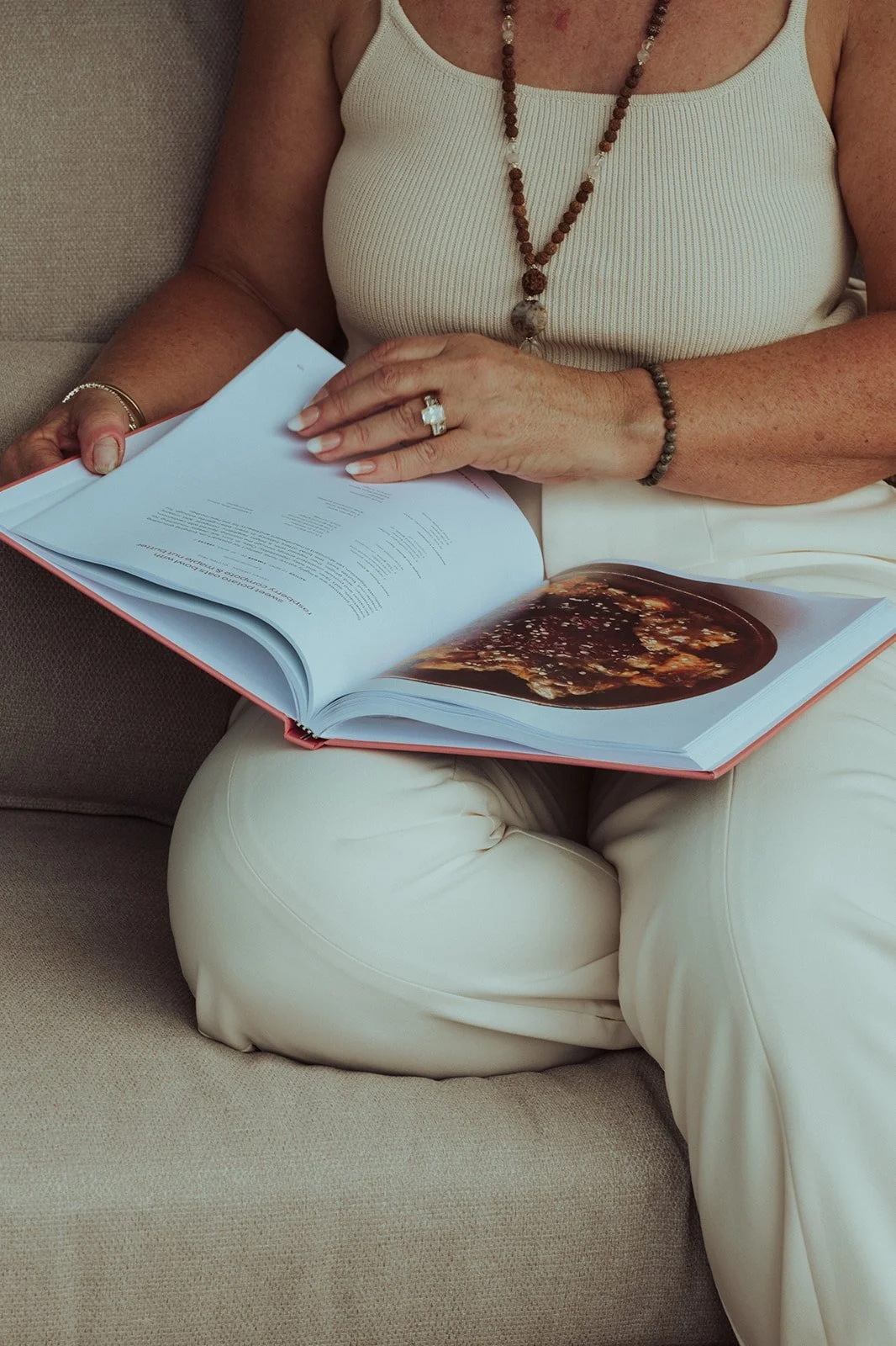 A woman wearing a white tank top and white pants sits on a beige couch, reading a large book with a picture of a dark, bubbling substance on the open pages. She is accessorized with a beaded necklace, a ring, and a bracelet.