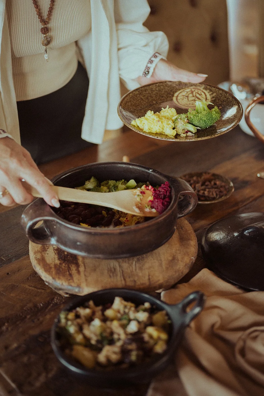 Person serving vegetarian dish from a black pot at a rustic wooden table, with various bowls of food including broccoli and scrambled eggs.
