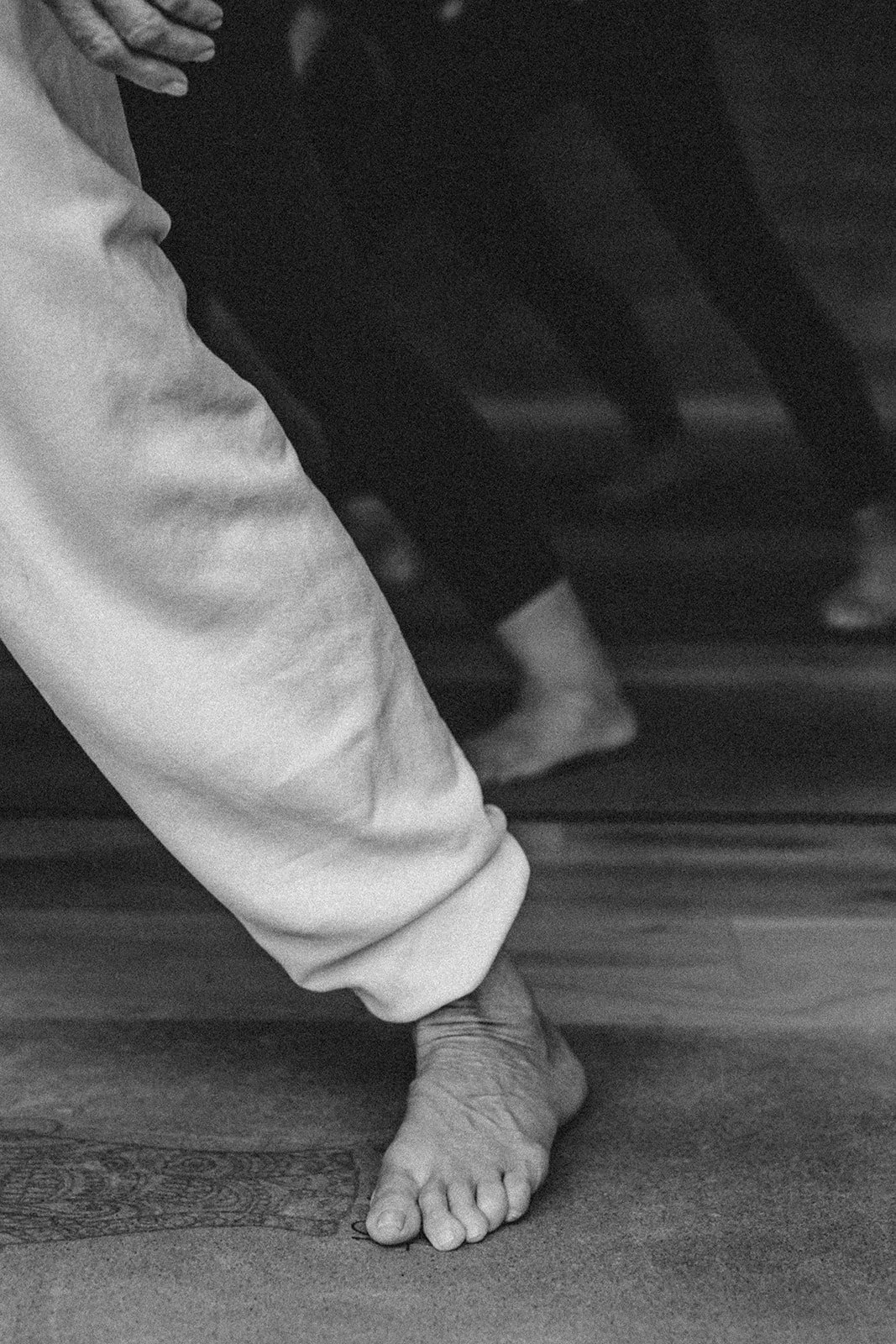 Close-up of a person with a hand on a knee, standing barefoot on a wooden floor, wearing light-colored pants and a dark top.