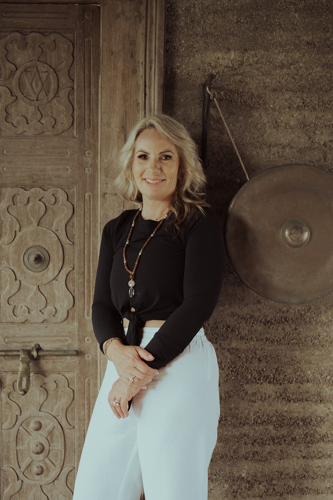 A woman with blonde hair and a black long-sleeved top, wearing white pants and a necklace, standing in front of a wooden door and a large gong mounted on a wall.