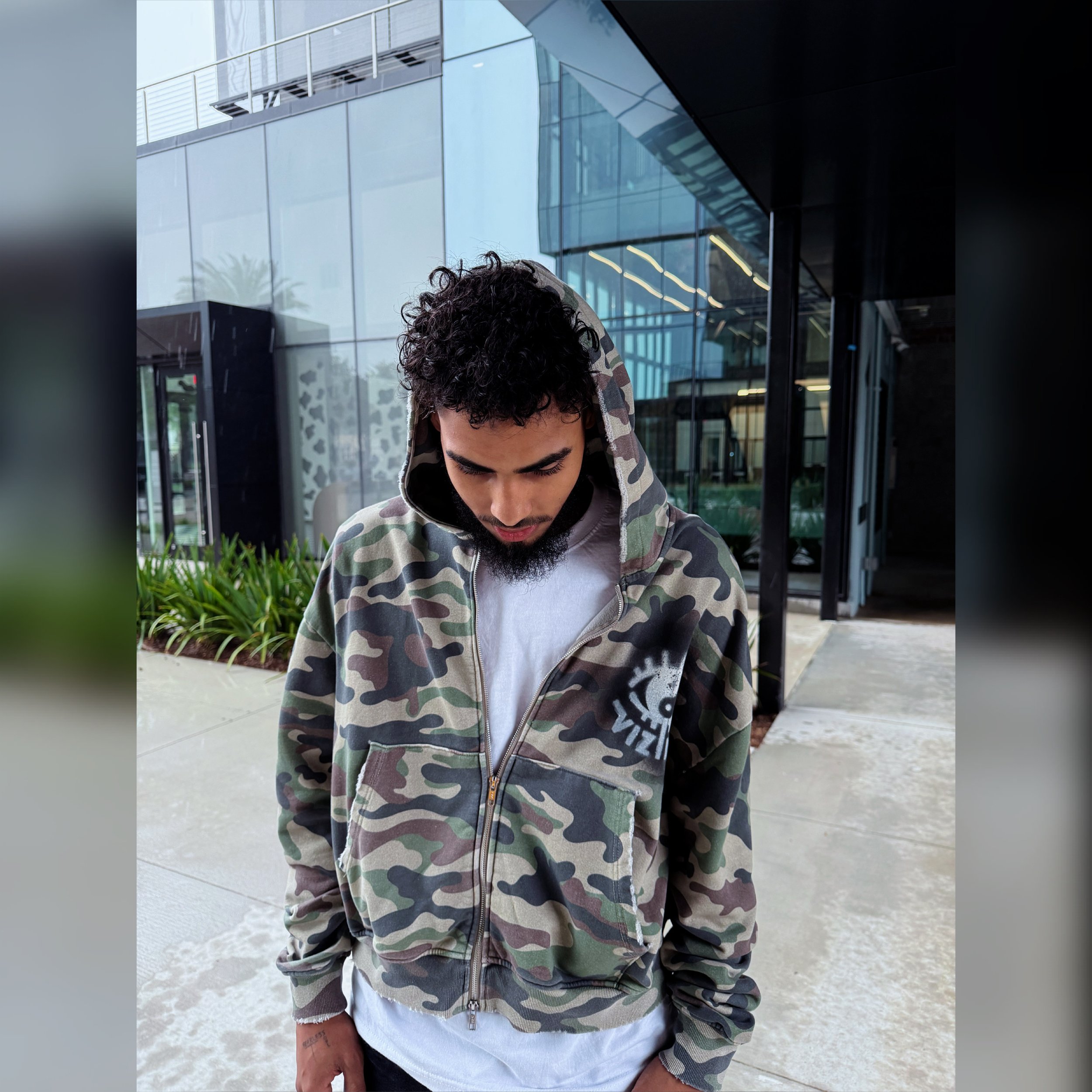 Close-up of a young man with curly hair and a beard, looking downward, wearing a camouflage hoodie and a white T-shirt, standing outside near a modern glass building with reflections and greenery.