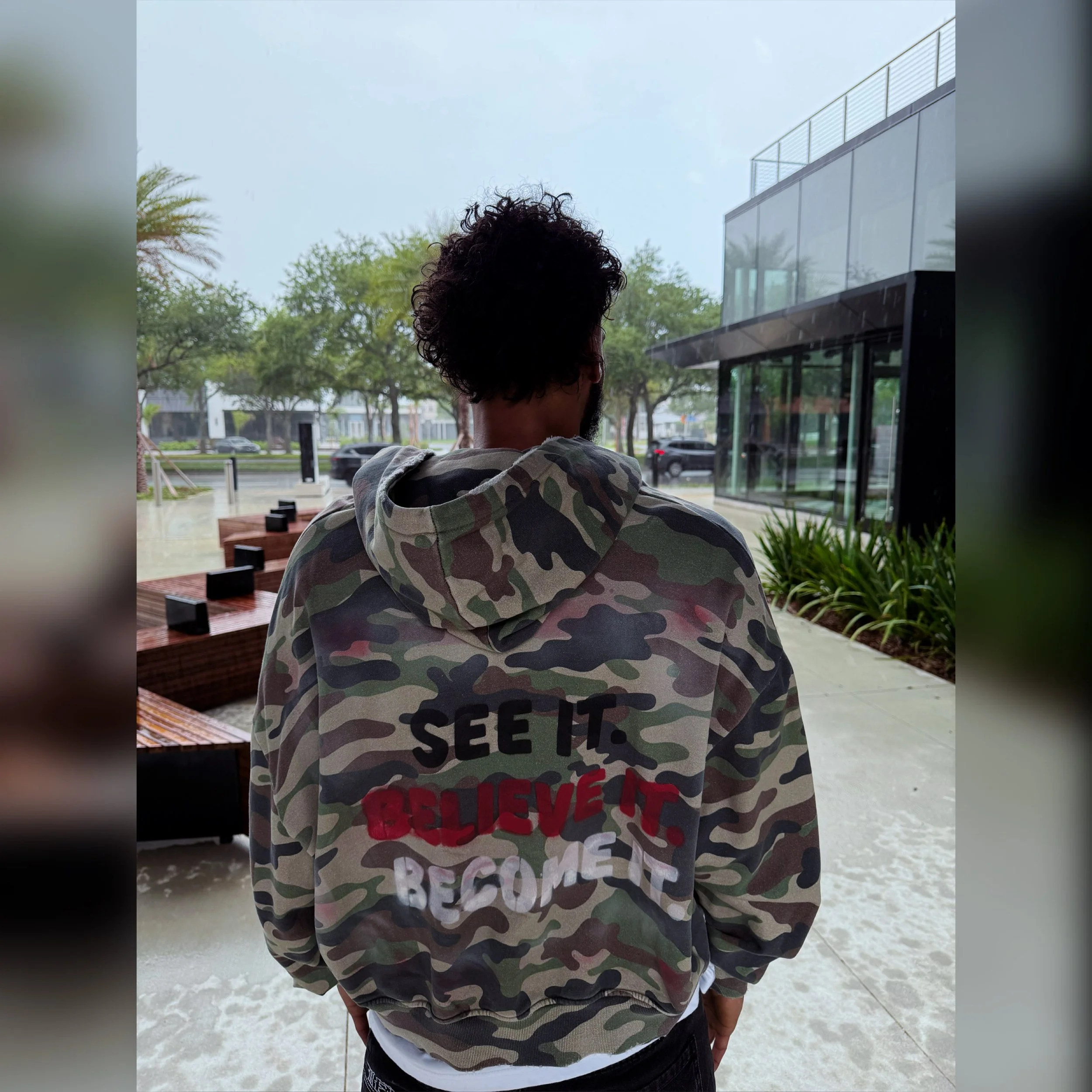 Person with dark curly hair wearing a camouflage hoodie, standing outdoors near modern glass building and benches, with trees and parked cars in the background.