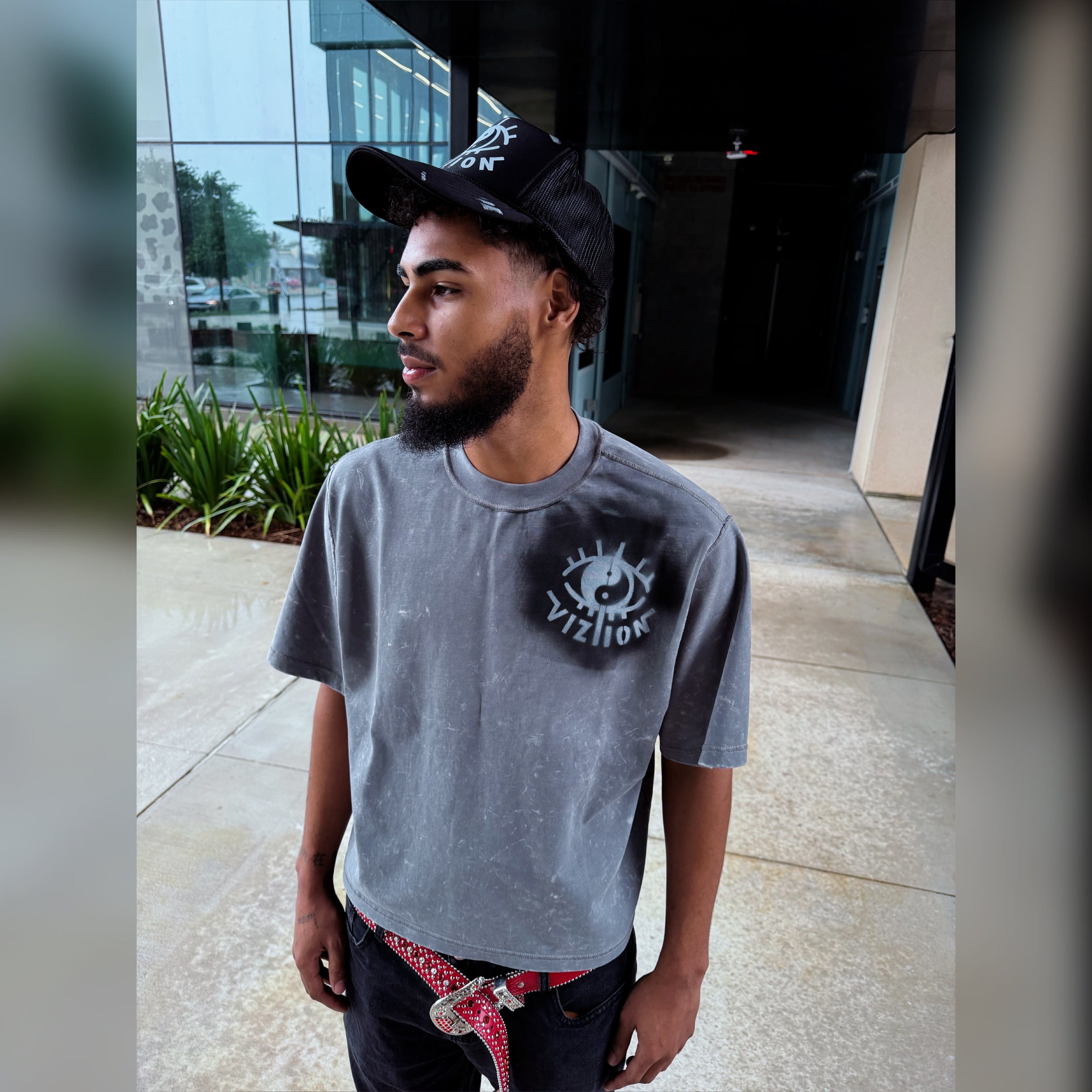 Close-up of a young man with a beard wearing a gray T-shirt, a black baseball cap, and a red belt with white polka dots, standing outside near a modern glass building with reflections of trees and cars.