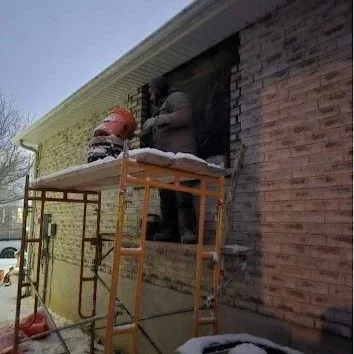 Two construction workers are on scaffolding outside a brick building, working on installing or repairing a window. Snow is visible on the ground and on the scaffolding.