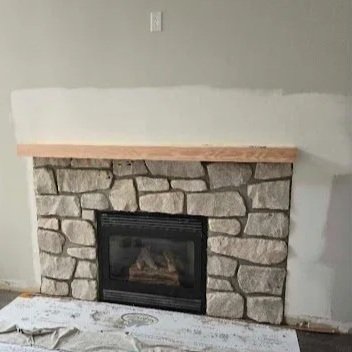 A stone fireplace with a wooden mantel against a gray wall, with an electrical outlet above it.