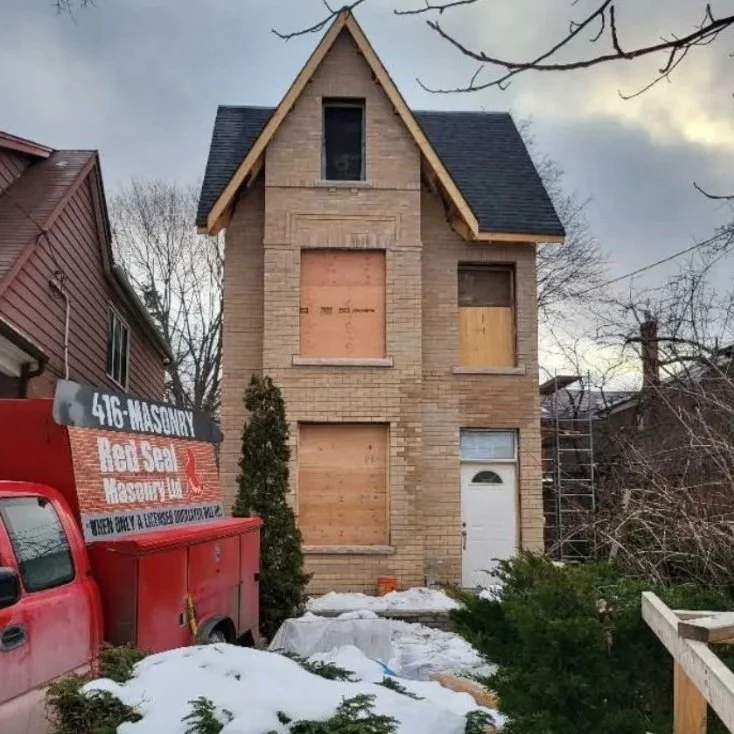 A three-story brick house under construction with boarded-up windows and an unfinished roof, surrounded by snow and trees, with a red construction truck parked in front.