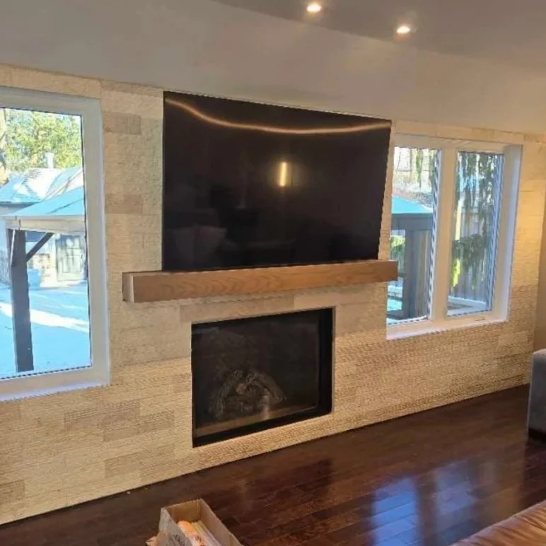 Living room wall with a flat-screen TV above a wooden mantel, a fireplace below, and two windows on either side showing an outdoor patio area.