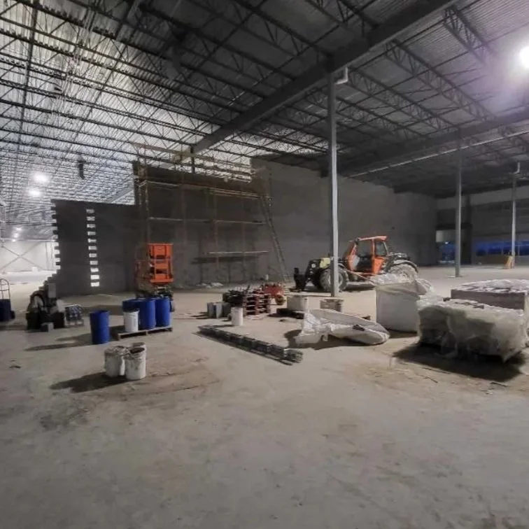 Interior of a large warehouse or construction site under construction, with construction equipment, materials, and scaffolding.