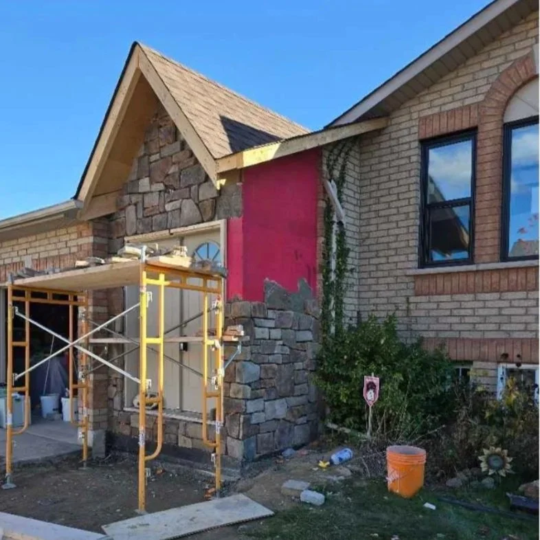 A house under construction showing brick and stone exterior with scaffolding in front and construction materials nearby.