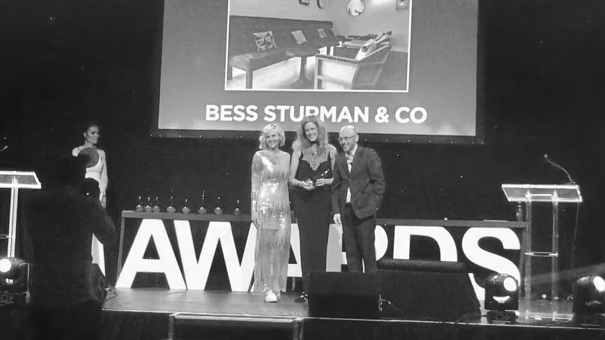 Three people standing on a stage at an awards ceremony, with a large sign reading "AWARDS" behind them and a projection screen above showing an interior room and the text "BESS STUPMAN & CO."