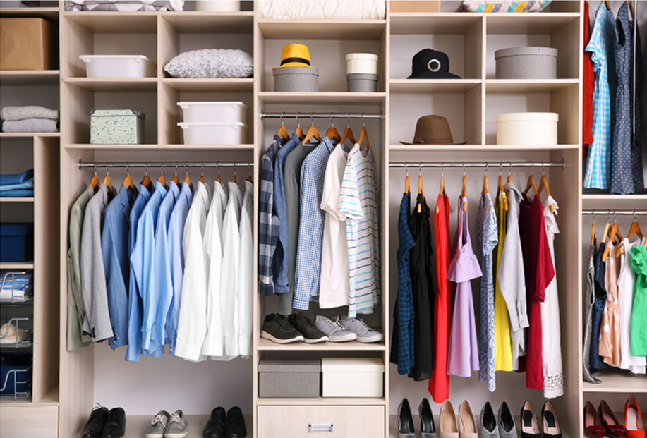 A closet with organized clothing, shoes, and storage baskets, featuring hanging shirts, pants, and dresses, with hats and boxes on shelves.