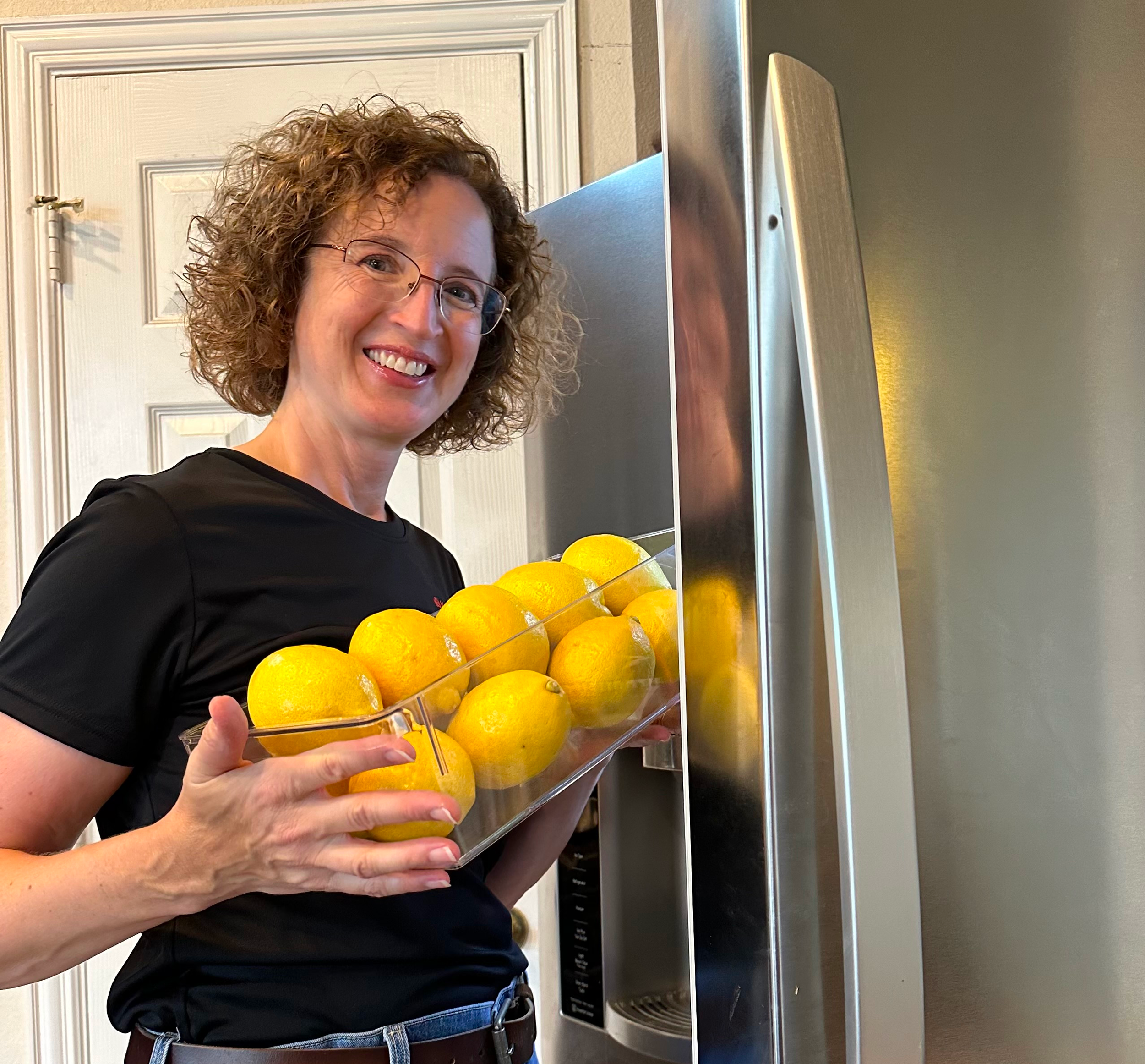 A woman with curly hair and glasses smiling while holding a clear tray of yellow lemons in front of a microwave oven in a kitchen.