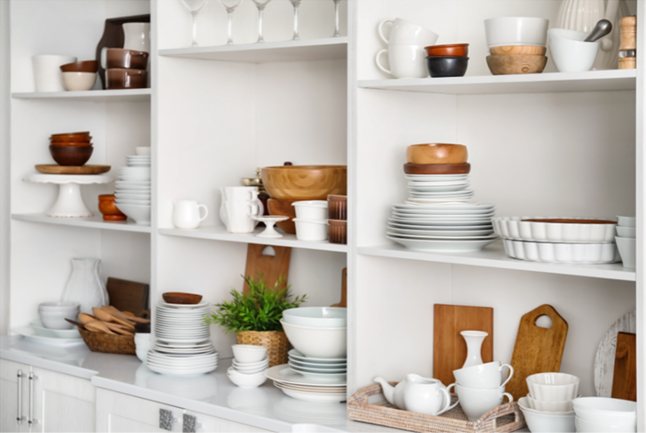 Open kitchen shelves filled with white, brown, and wooden dishes, bowls, and kitchenware, with some greenery and cutting boards.